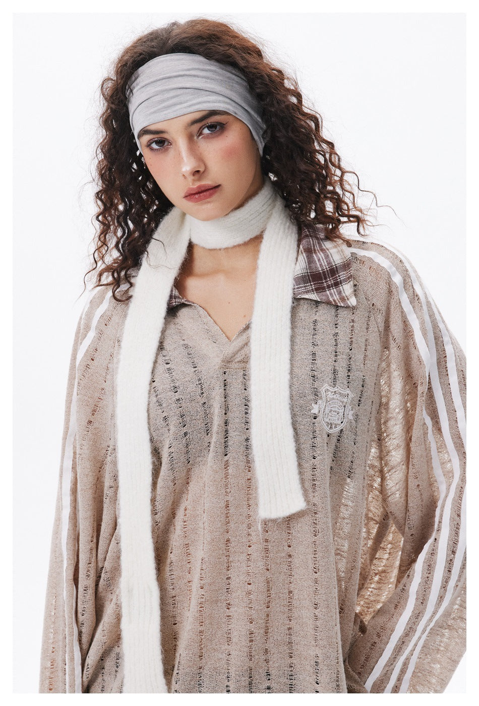 Close view of a model wearing a beige open-knit pullover with white sleeve stripes, plaid collar, scarf, and gray headband.