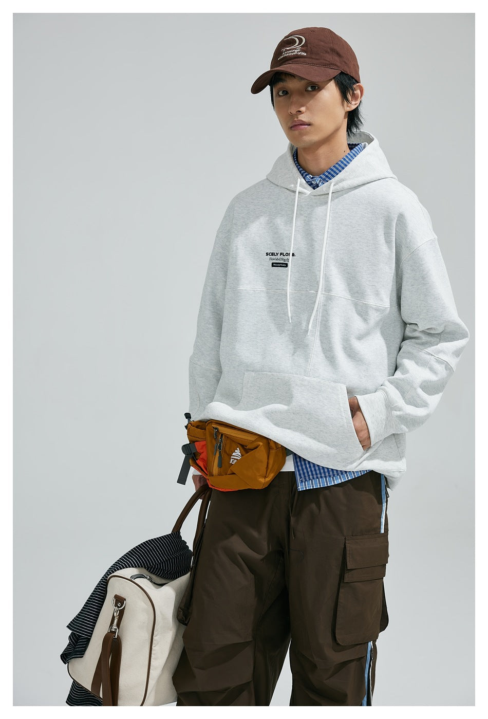 Close-up of a model in a light gray hoodie with chest print, hand in pocket, with waist bag and brown cap
