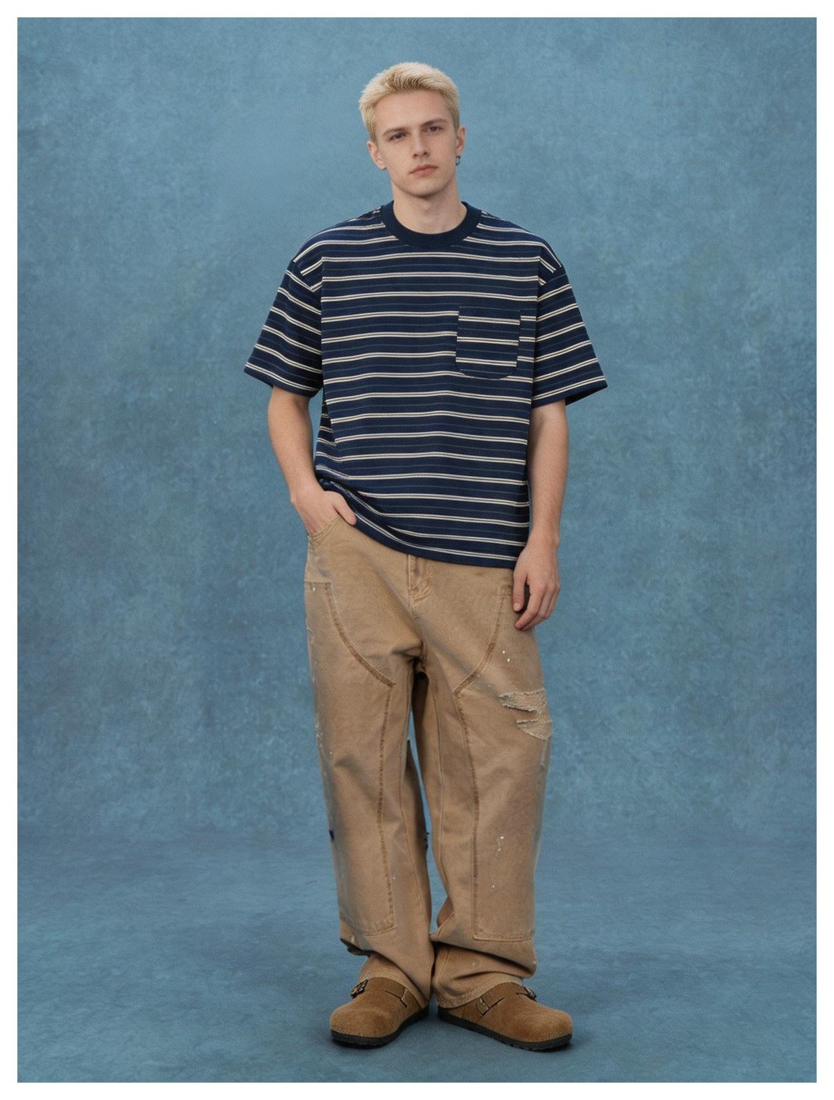 Man wearing navy striped pocket T-shirt with khaki wide-leg work pants and brown clogs against a blue backdrop