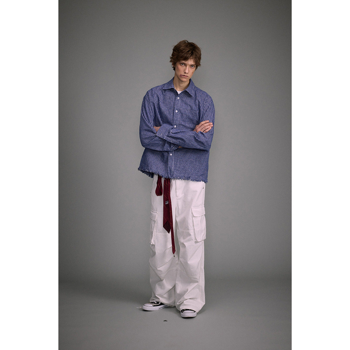 Model in a blue small-check button-up shirt with a frayed hem, white cargo pants, and a burgundy tie.