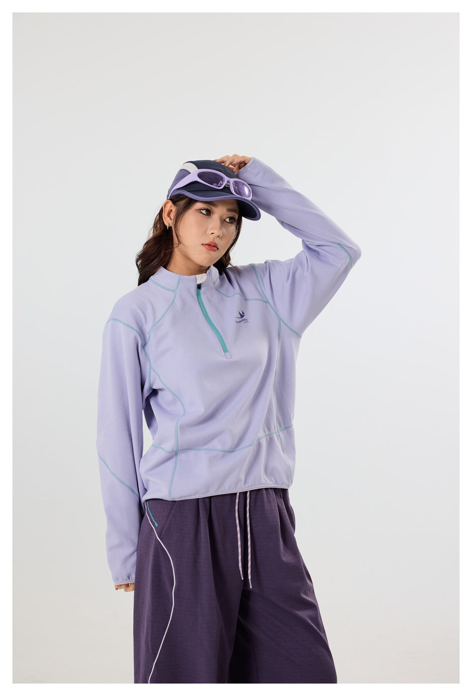 A woman models a lavender quarter-zip pullover with teal seam accents and dark purple pants, shown from the thighs up.
