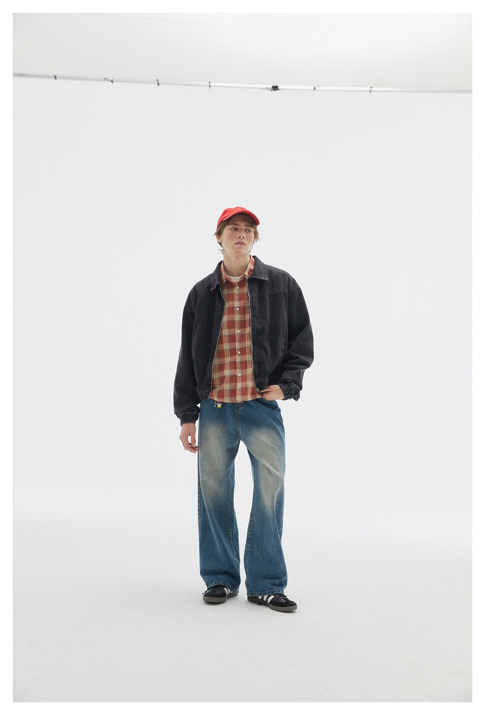 Person standing in a dark gray denim zip jacket over a red plaid shirt, red cap and wide-leg jeans