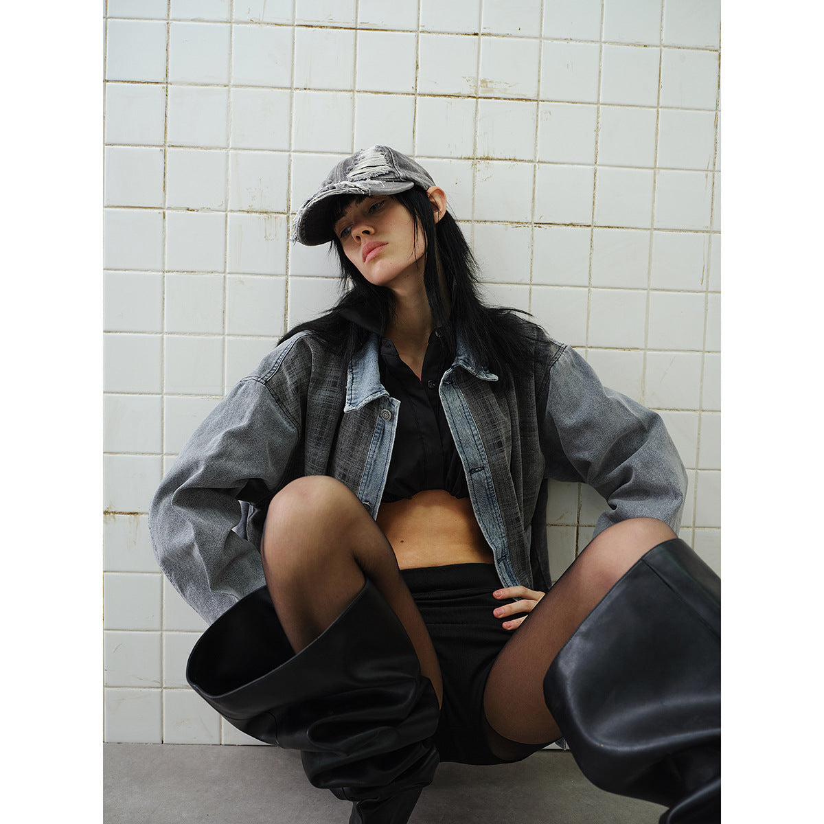 Seated model in an oversized charcoal plaid jacket with denim trim, black shorts, sheer tights, and tall black boots.