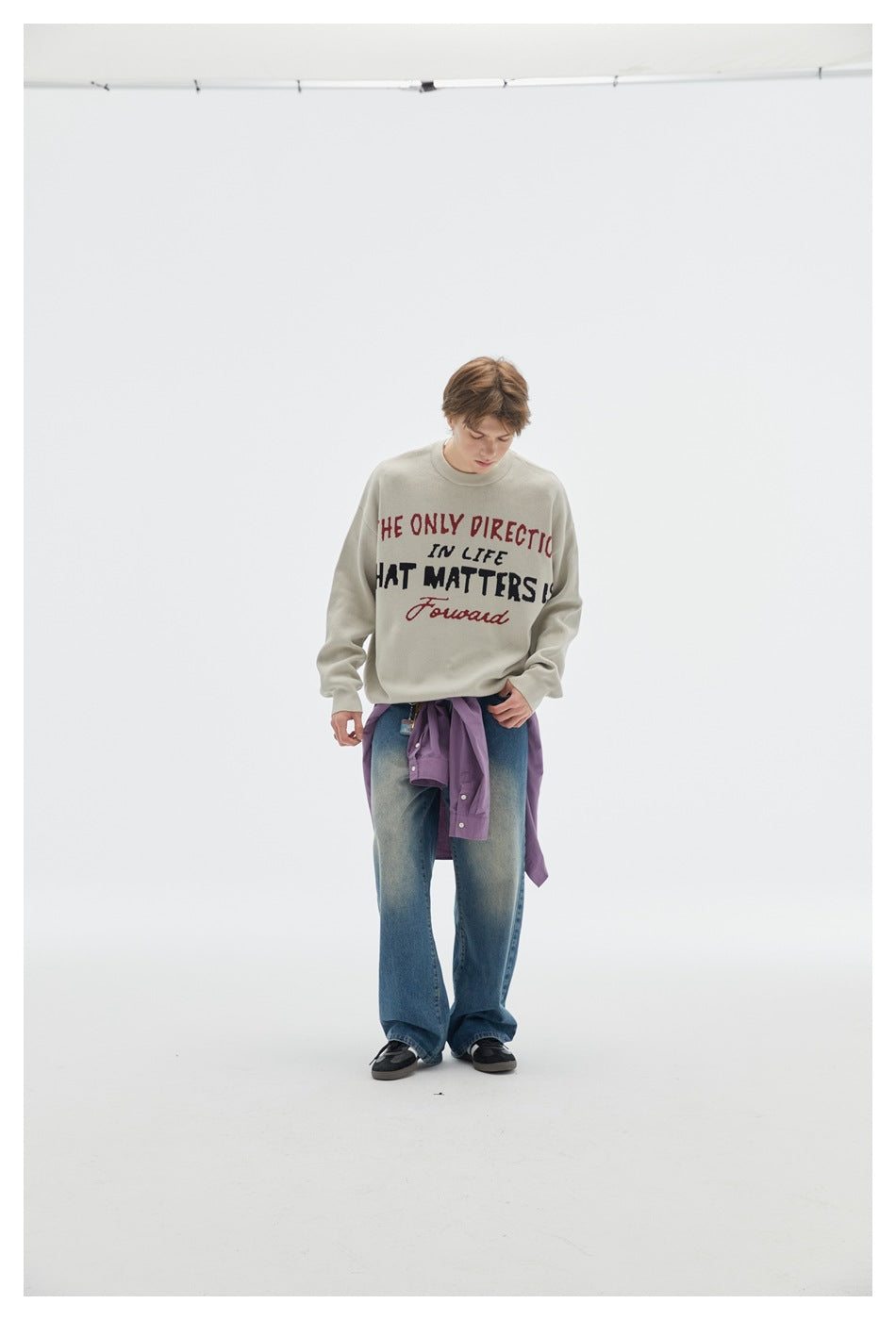 Person wearing a light beige slogan crewneck sweater with wide-leg jeans and a purple shirt tied at the waist