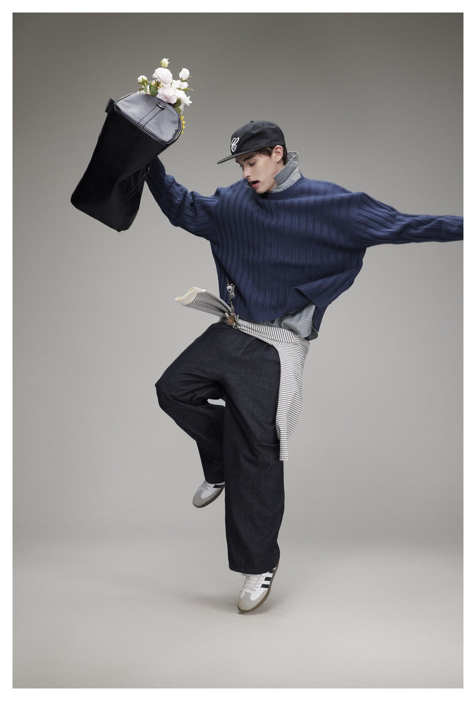 Model in a blue ribbed sweater and dark wide-leg jeans, posing with a black bag and wearing a black cap.