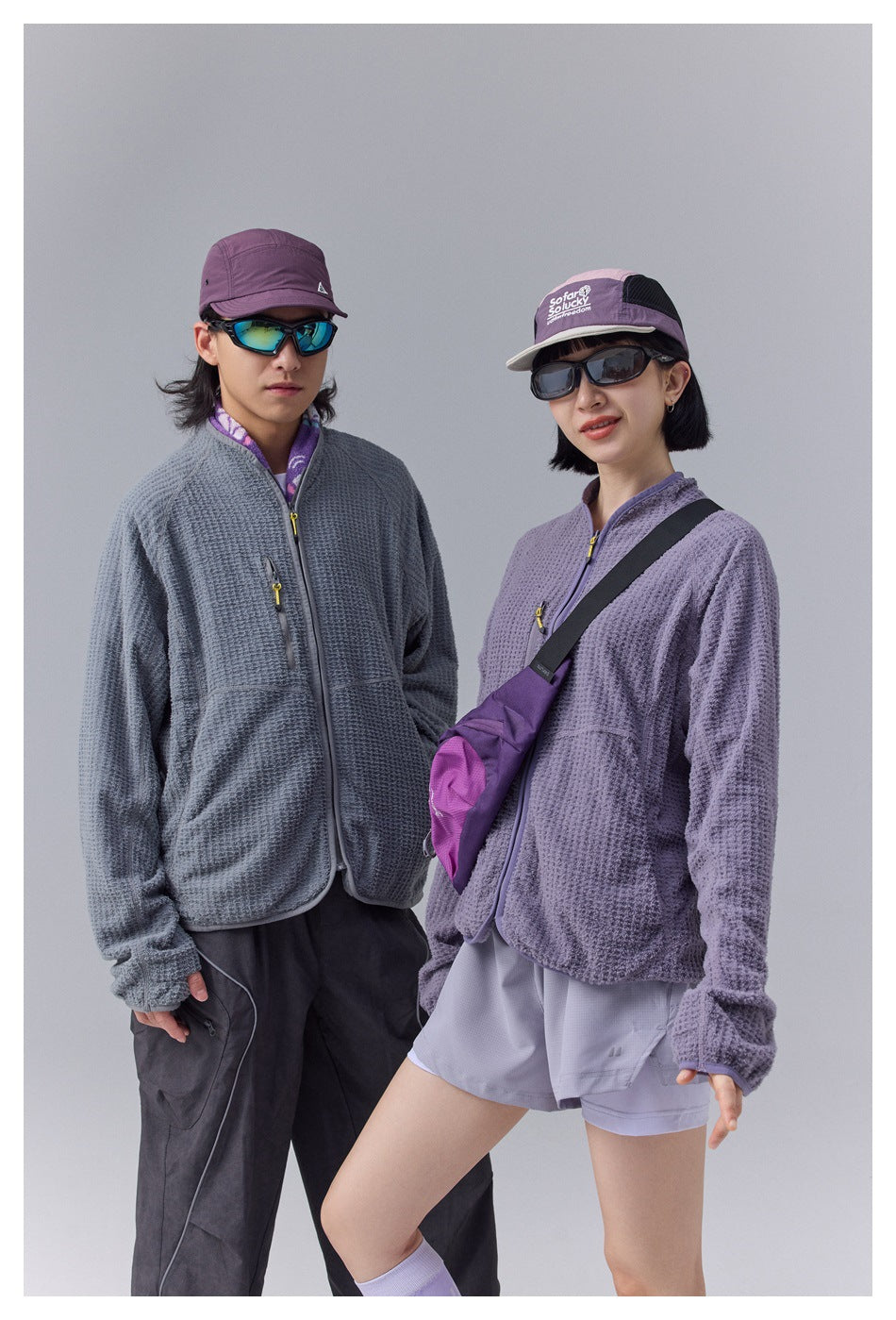 Two models wearing gray and purple textured zip fleece-style jackets, with caps, sunglasses and a crossbody bag, close-up