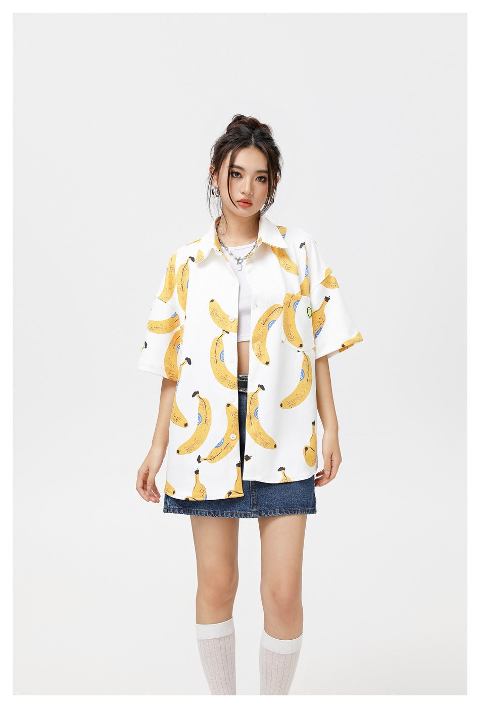 Front view of a model in an oversized white banana-print shirt with short sleeves, styled over a blue denim skirt.