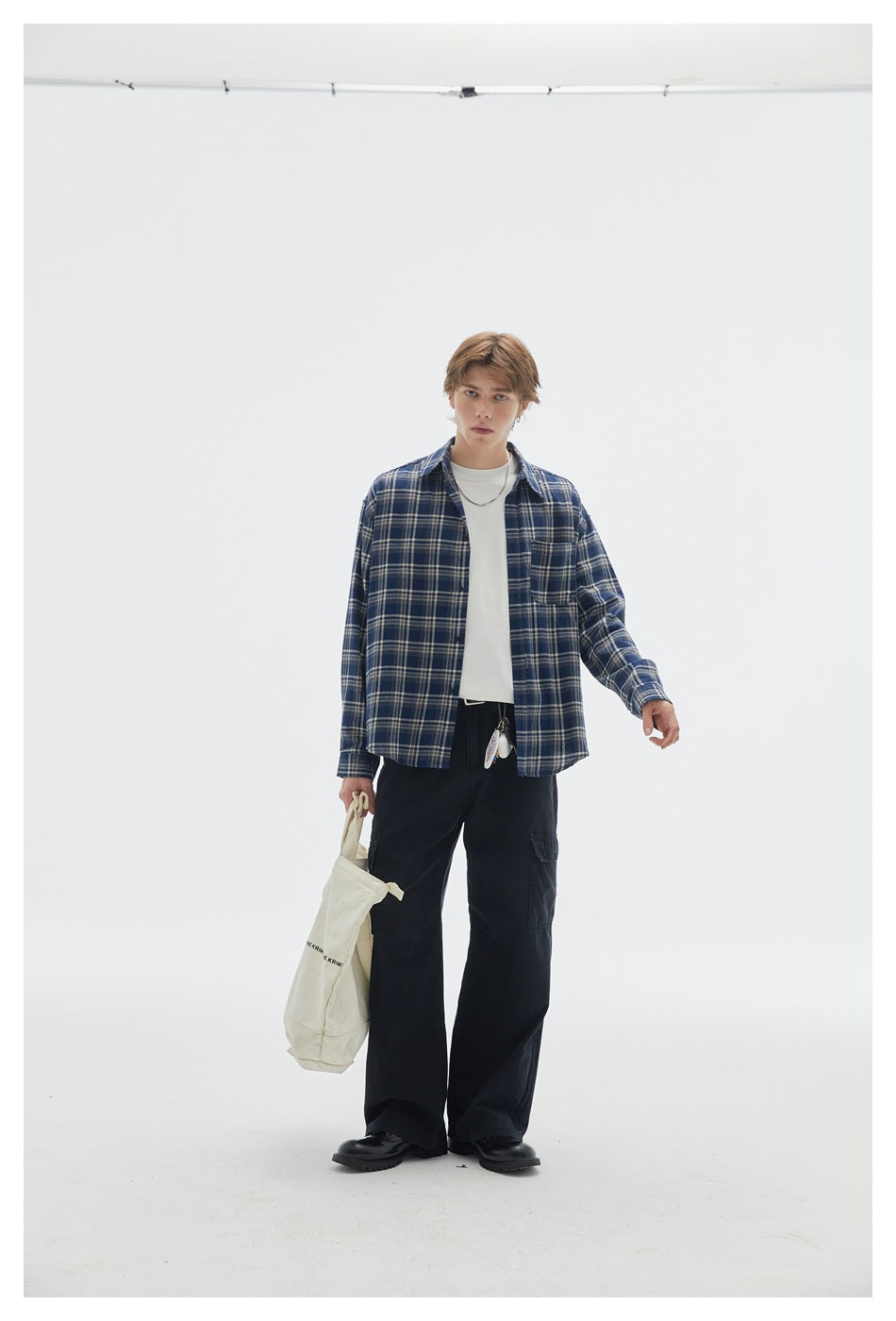 Model in black wide-leg cargo pants, blue plaid shirt and white tee, holding a cream canvas tote bag