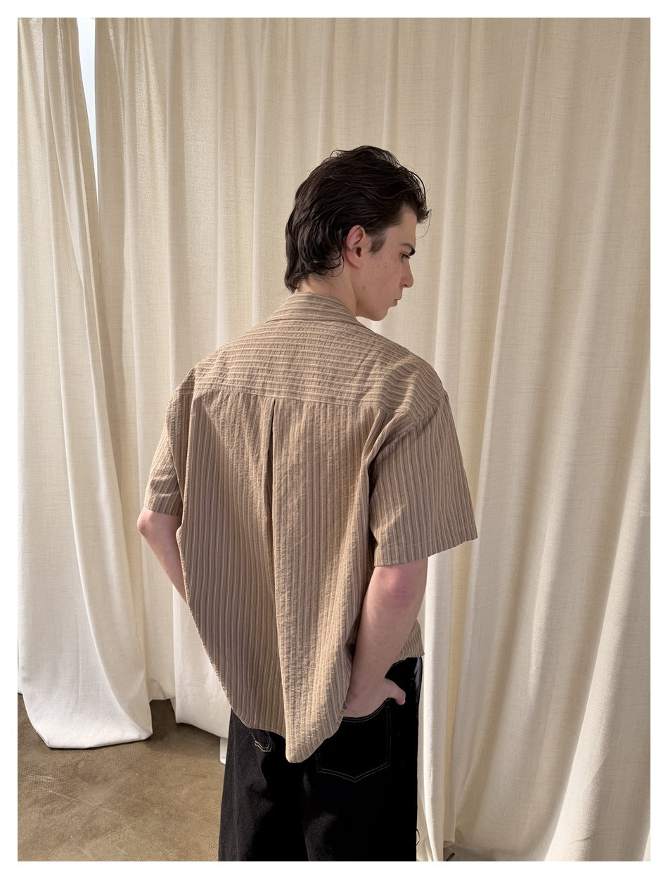 Three-quarter rear view of a model wearing a taupe striped short-sleeve shirt with a pleated back.