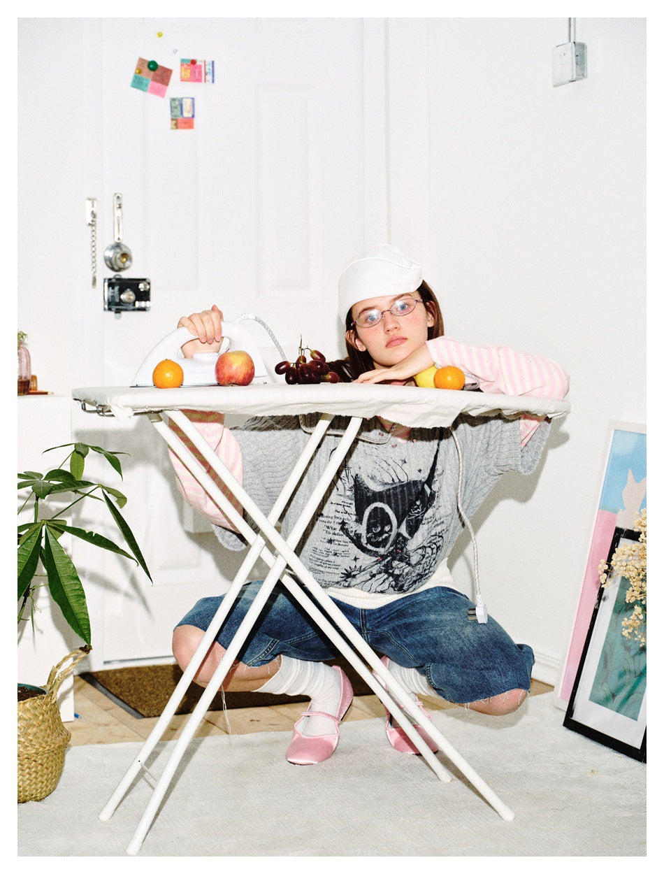 Model wearing a gray graphic top over pink stripes and denim shorts, crouched behind an ironing board.