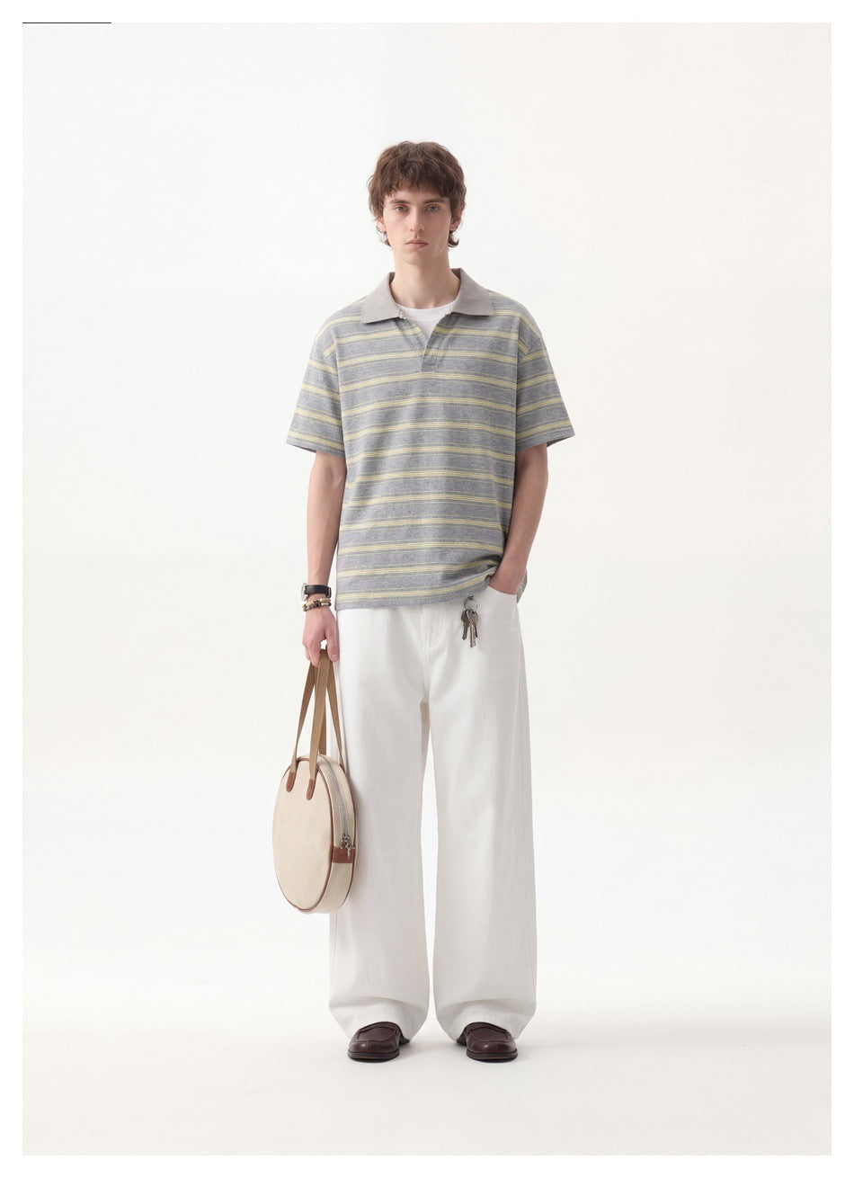 A man models a gray polo with thin yellow stripes and white wide-leg trousers, holding a round beige bag.