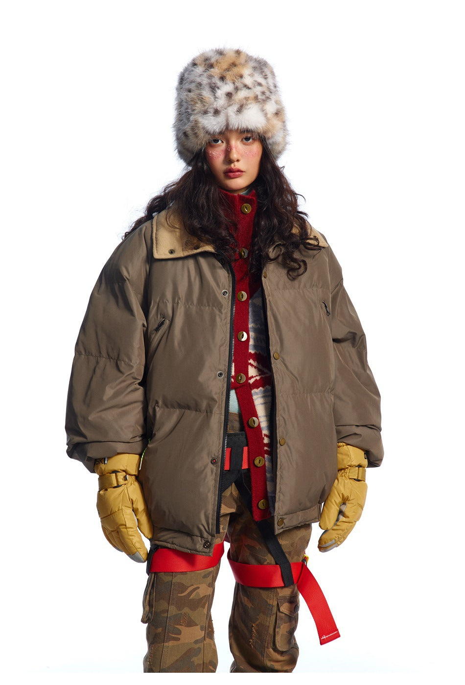 Front view of a model in a dark taupe oversized puffer jacket, red patterned cardigan, camouflage trousers and spotted fur