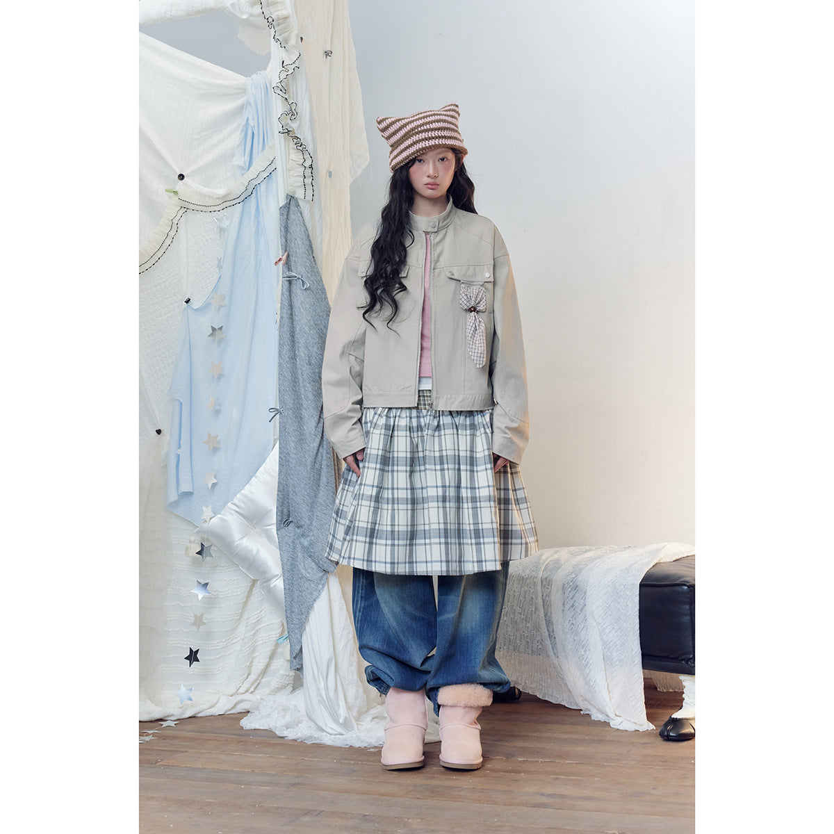 Model wearing a light gray jacket, pink top, plaid skirt over jeans, and striped knit hat.