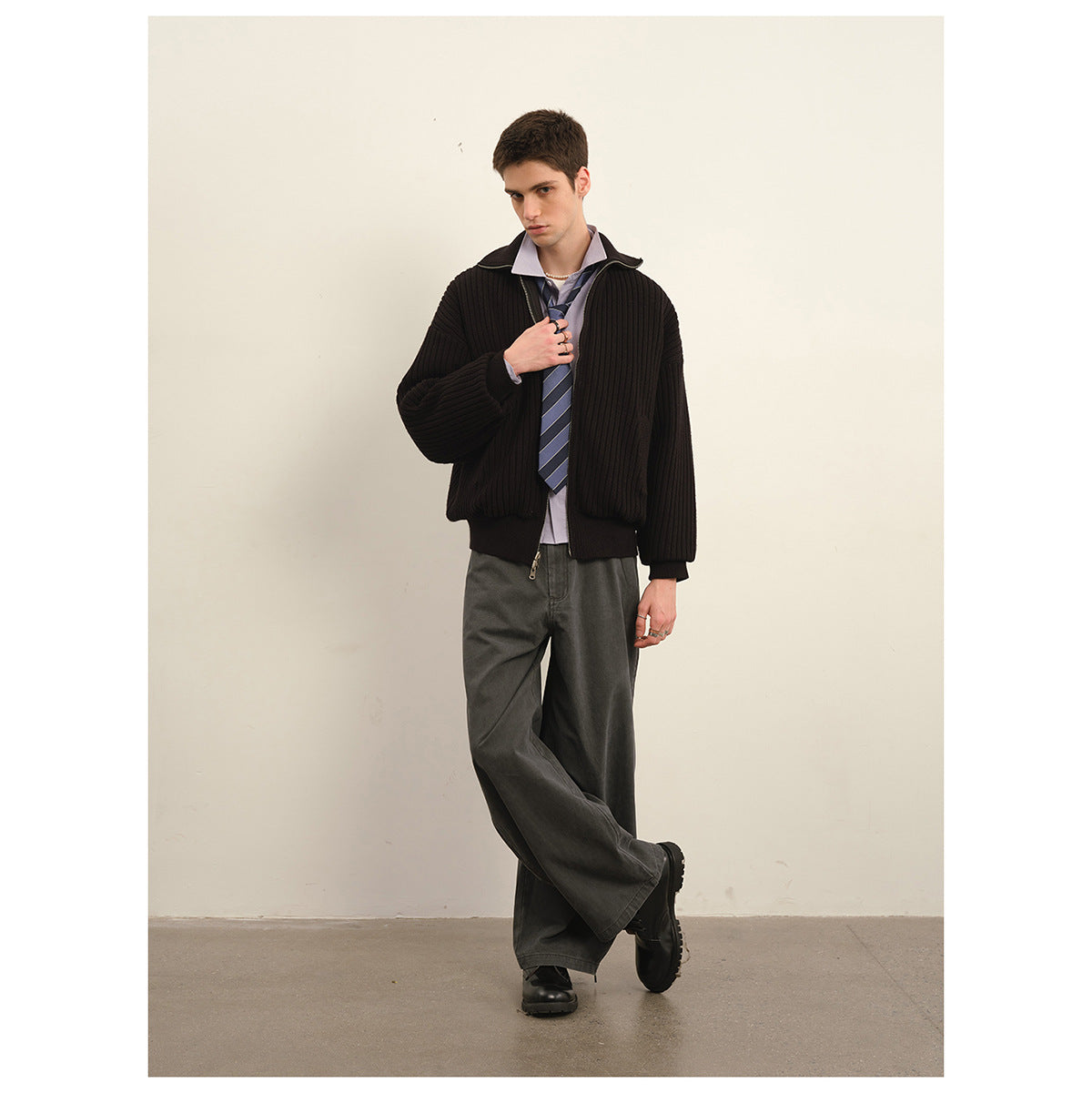 Model wearing a black ribbed zip jacket over a lavender shirt and striped tie with wide charcoal trousers.