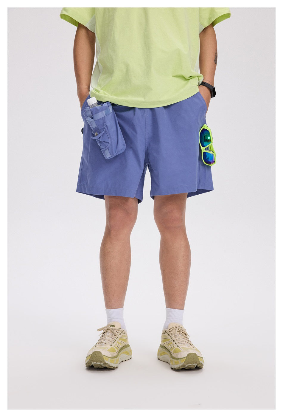 Front view of a model wearing periwinkle-blue shorts with utility pockets, a light lime T-shirt, and beige sneakers.