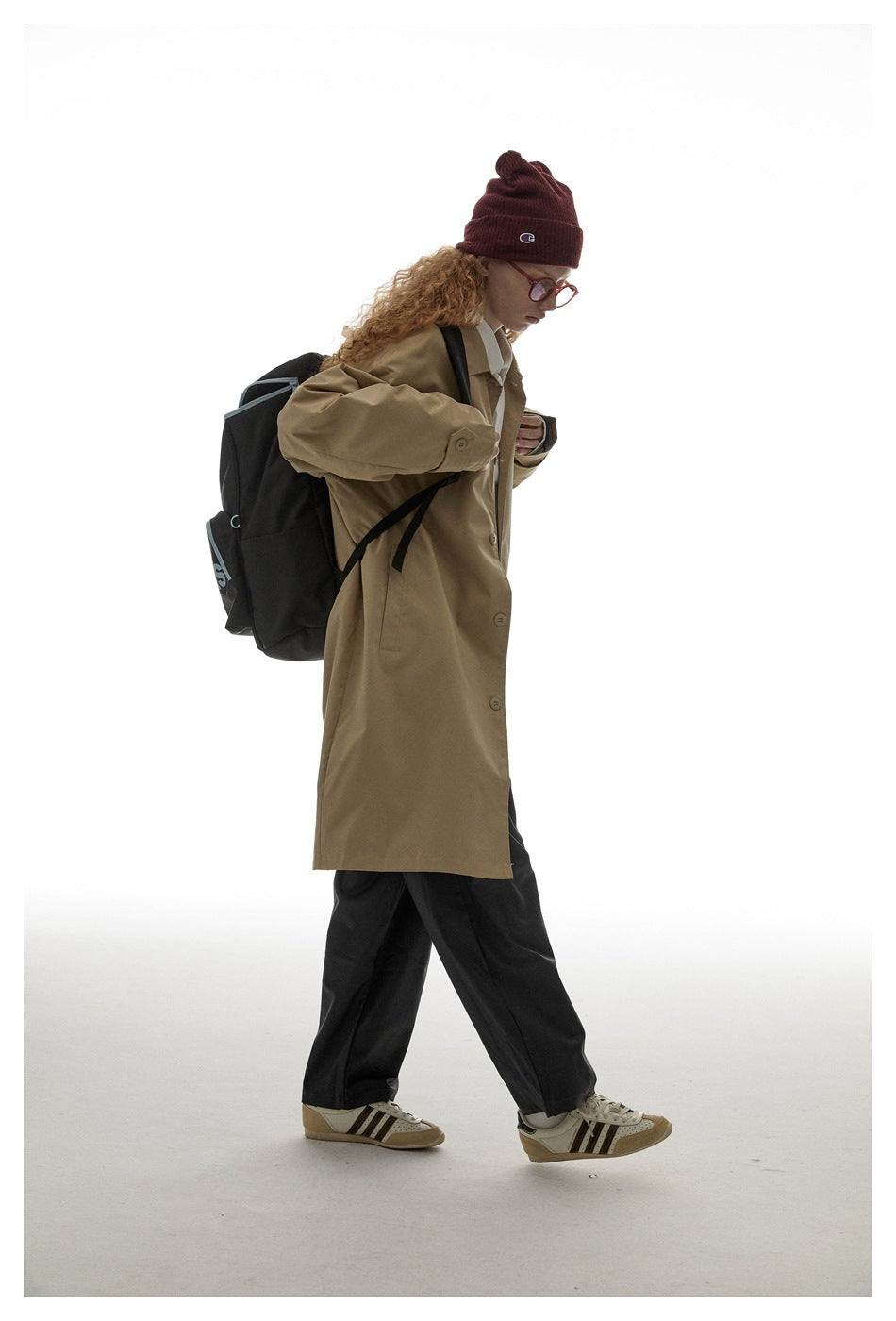 Side view of a model wearing a tan long coat with button cuffs and a black backpack.