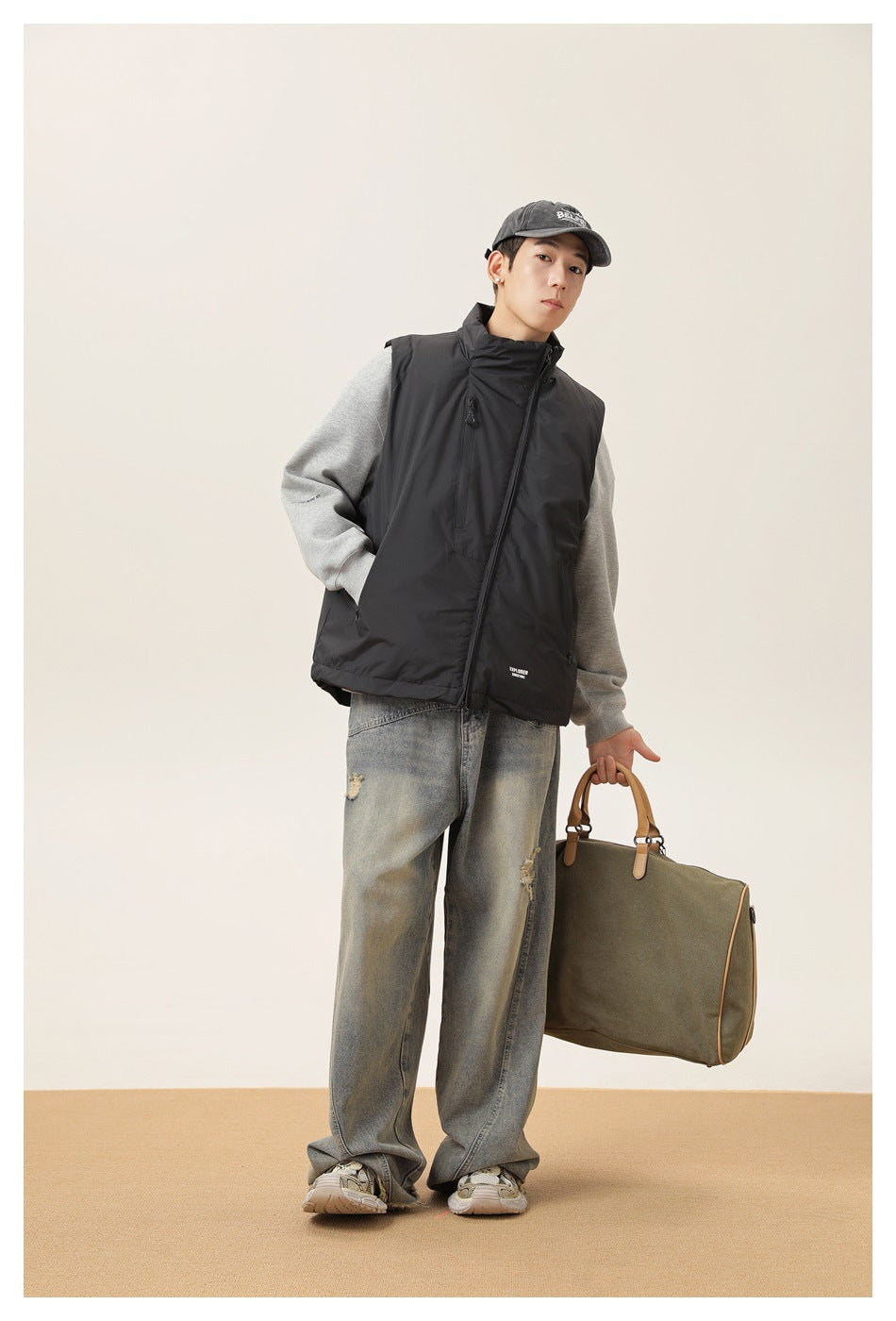 Model wearing a black high-collar sleeveless vest over a gray sweatshirt, with loose jeans and a cap.