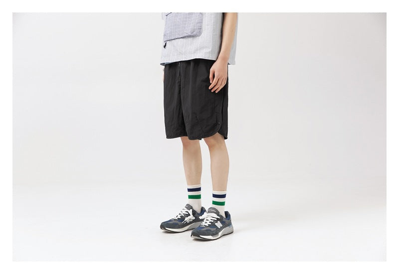 Side view of a model wearing black knee-length shorts with a light shirt and sneakers.