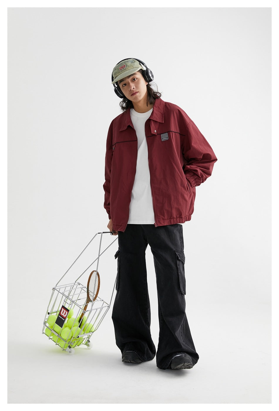 Full-length model wearing a burgundy zip jacket, black cargo pants and sage cap, with headphones and a tennis ball basket.