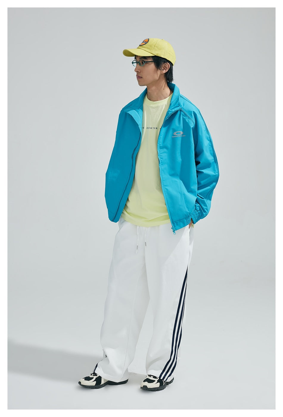 Person wearing an open sky blue stand-collar jacket with a yellow tee, white striped pants and a yellow cap