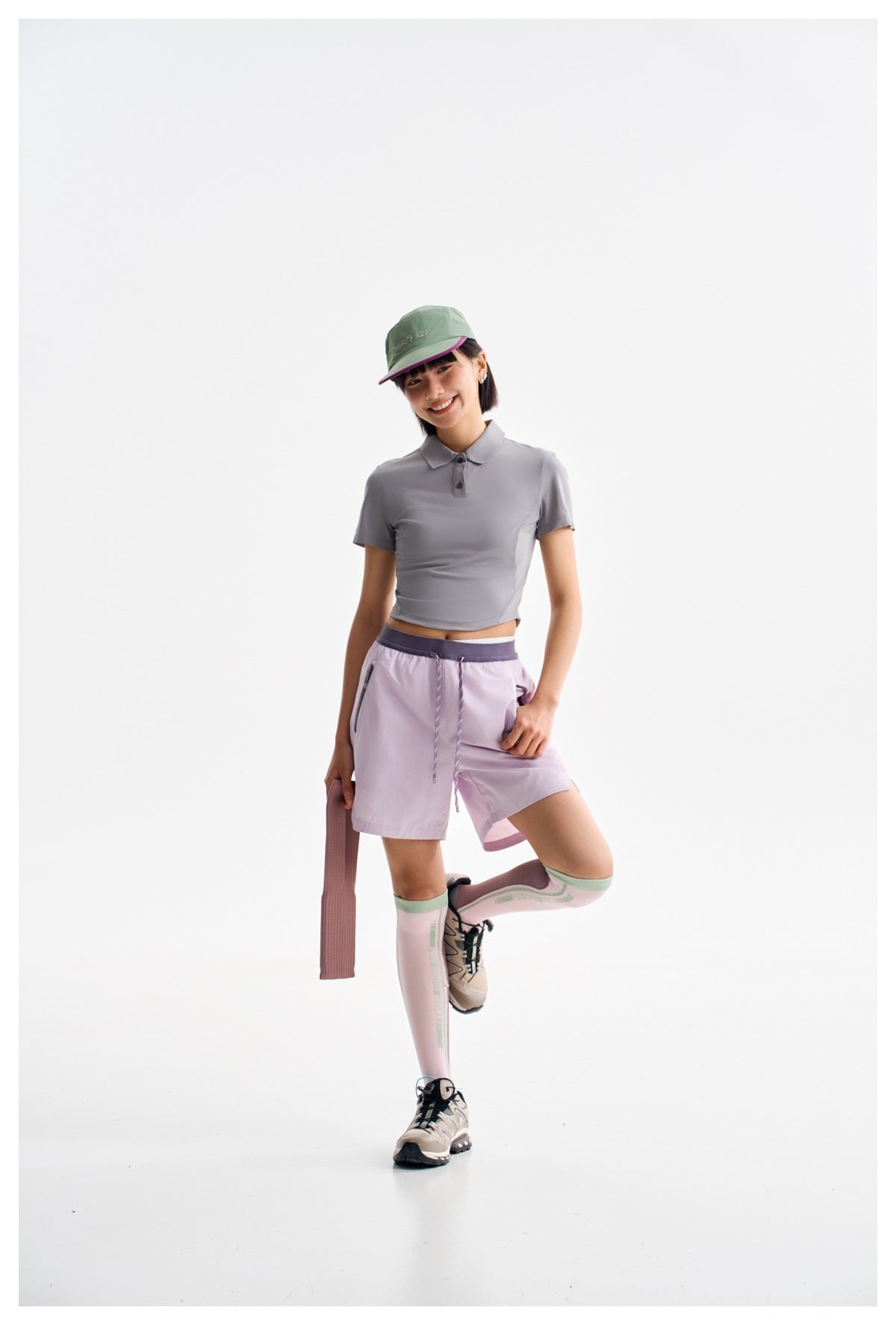 Model in a gray cropped polo and lilac drawstring shorts, with a mint cap and pink knee-high socks.