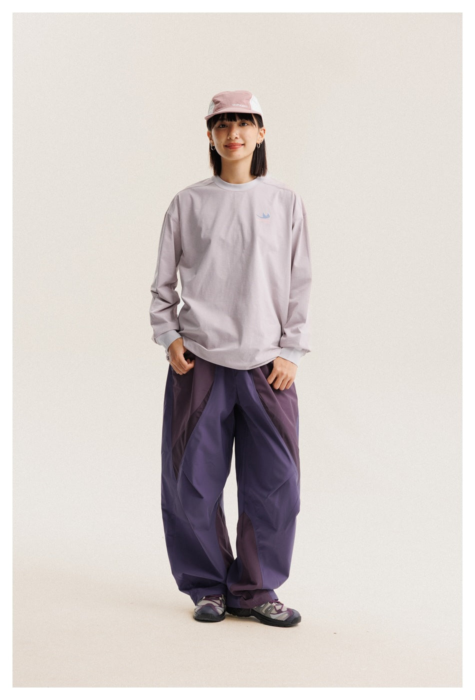 Full-length model wearing a pale lilac long-sleeve shirt, loose paneled purple pants, and a pink cap.