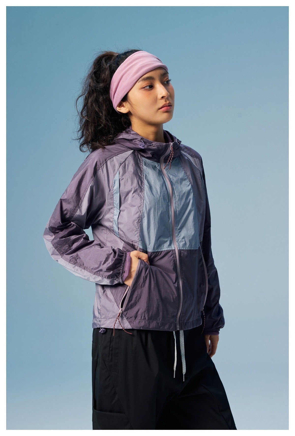 Model wearing a purple and pale blue color-block hooded jacket, shown from the front with its zip pockets visible.