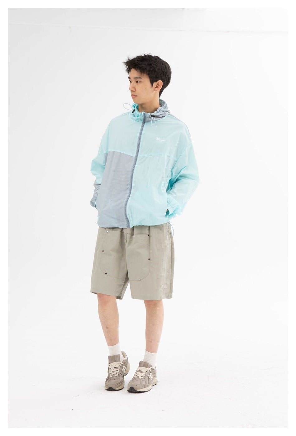 Model standing in gray knee-length shorts with patch pockets, a light blue and gray zip jacket and sneakers