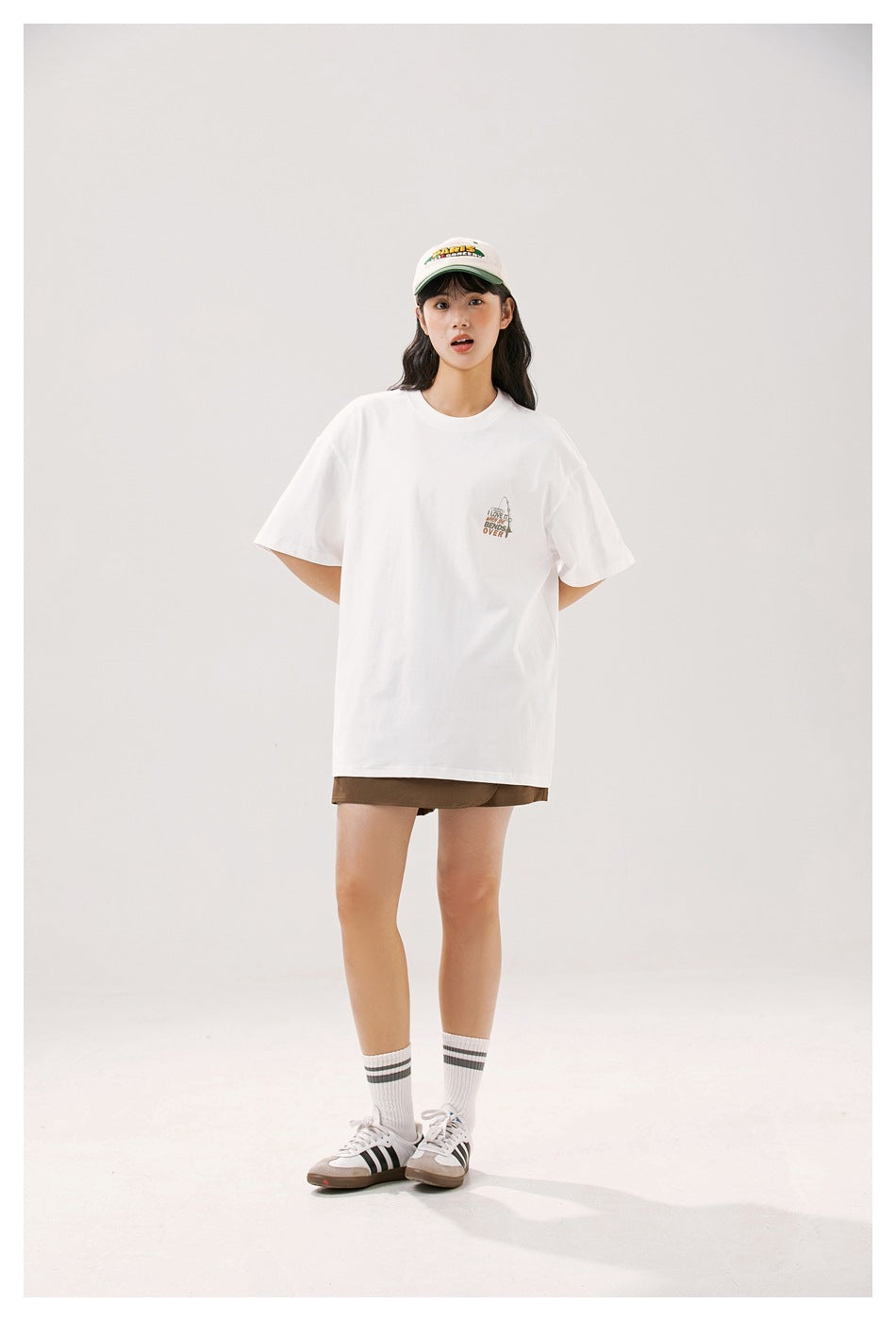 Model wearing a white oversized T-shirt with a small chest print, brown shorts, a cap and sneakers