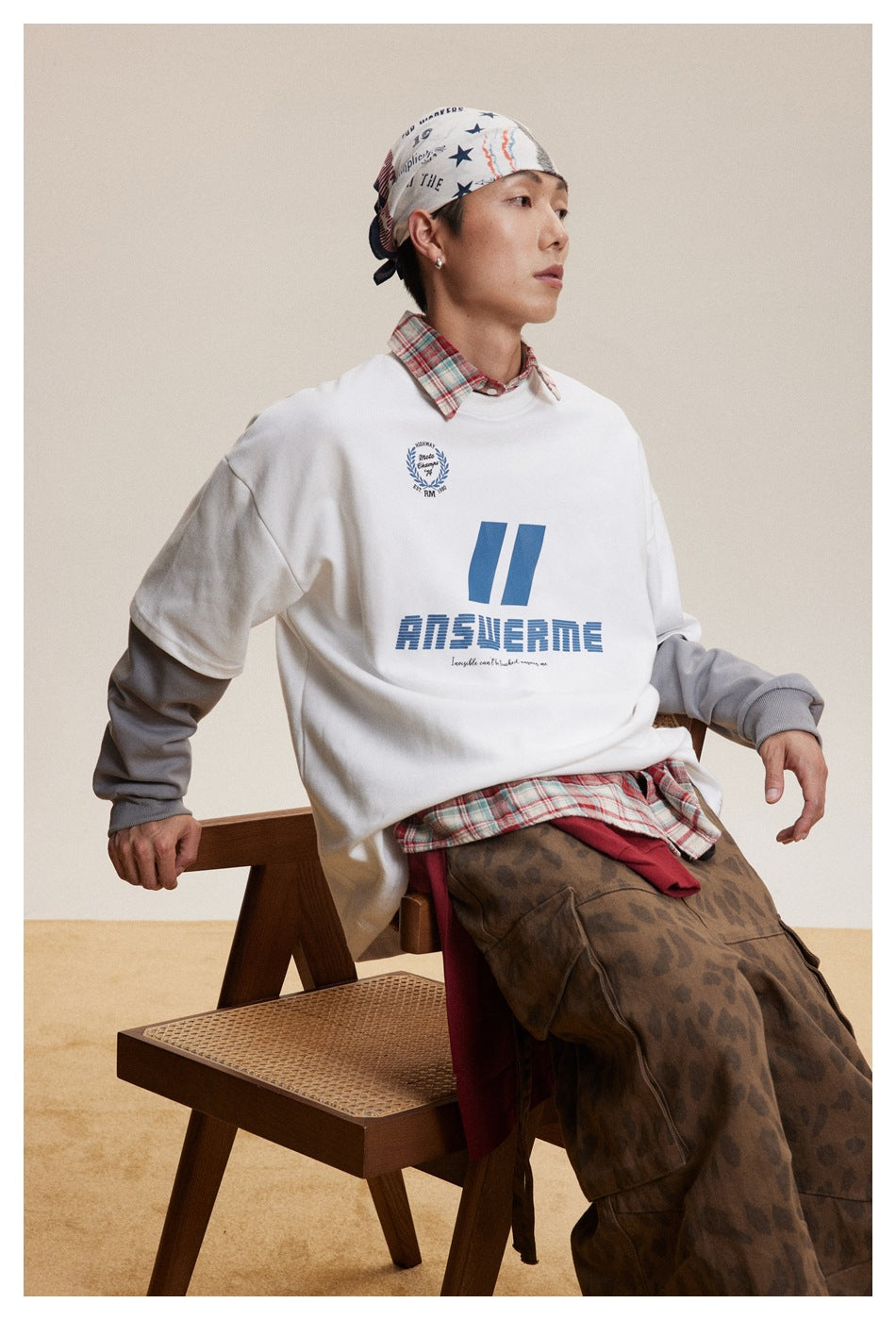 Model seated in a white graphic sweatshirt layered over gray sleeves, with a plaid shirt underneath.