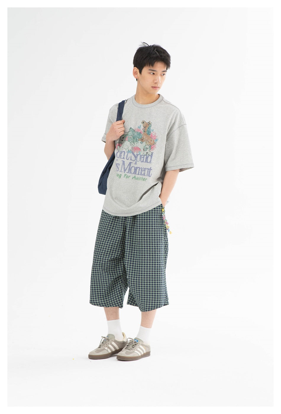 Model in a heather gray graphic T-shirt with plaid cropped pants and sneakers, a denim bag over one shoulder