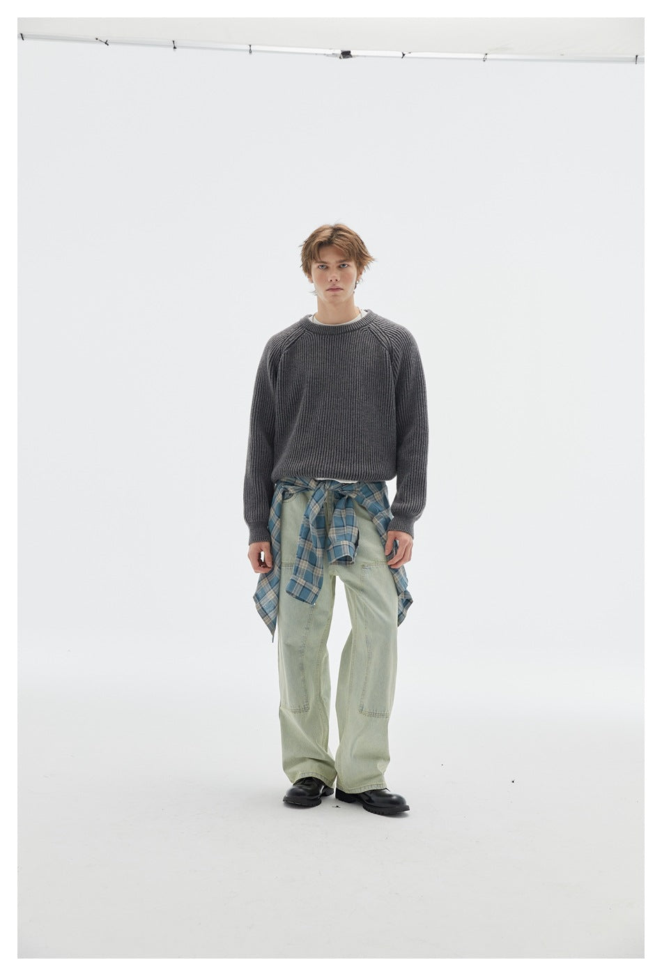 Full-length front view of a model in a charcoal ribbed crewneck sweater, pale green jeans, and a blue plaid shirt tied at the