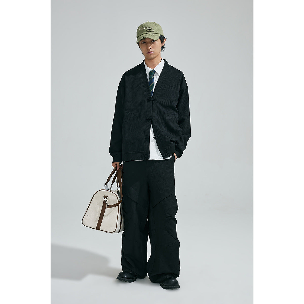 Model wearing a black frog-button cardigan and wide-leg trousers with a white shirt and striped tie, carrying a duffel bag.