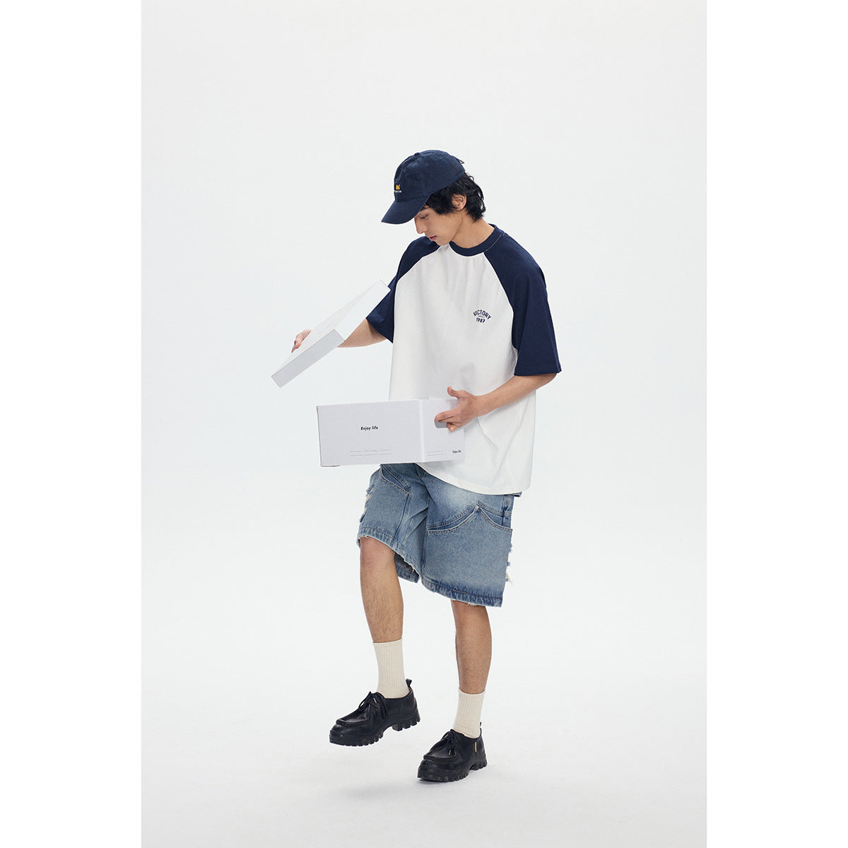 Model wearing a white raglan T-shirt with navy sleeves and light blue denim shorts, holding a box.