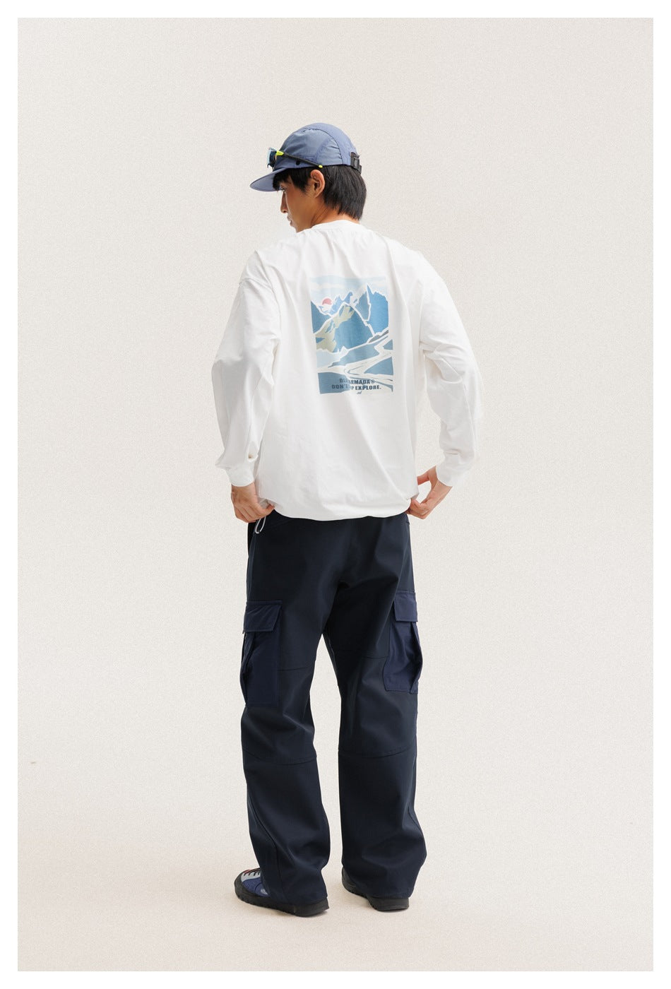 Rear view of a model wearing a white long-sleeve shirt with a blue mountain graphic and navy cargo pants.