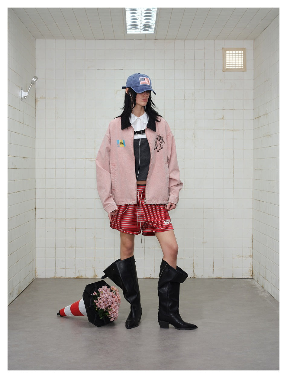 Model wearing a dusty pink graphic jacket, red striped shorts, blue cap, and tall black boots.