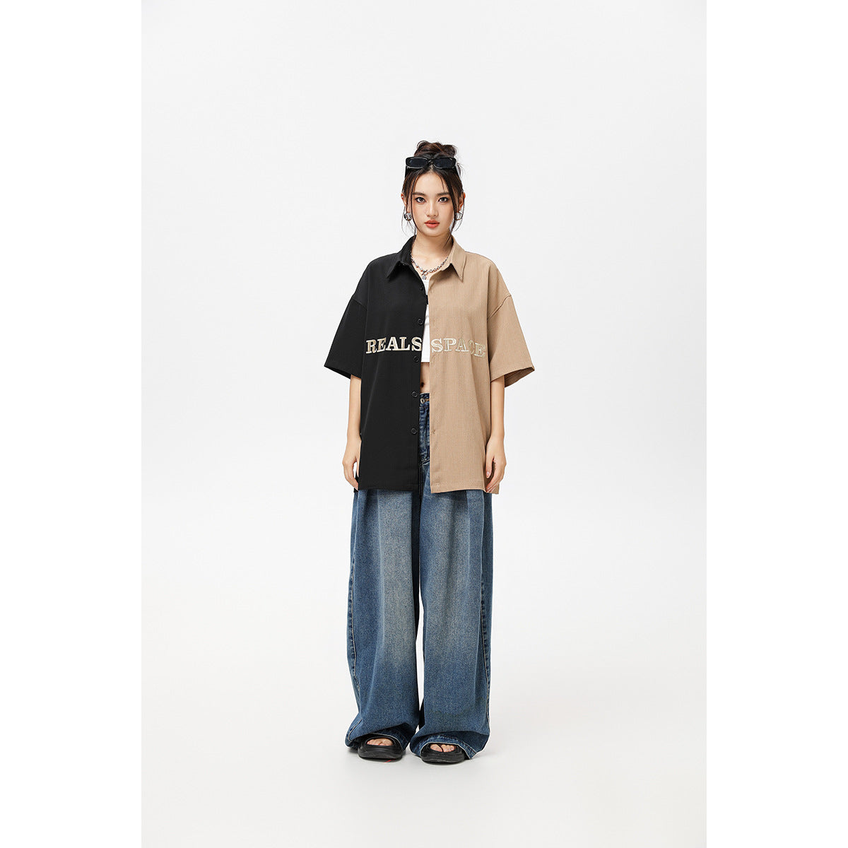 Full-length model wearing an oversized short-sleeve shirt split black and tan with embroidered lettering and wide-leg jeans.