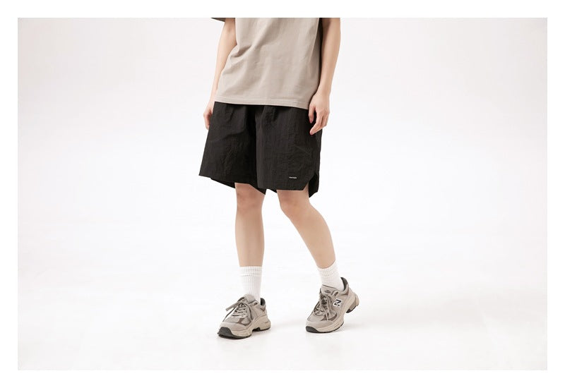Cropped front view of a model wearing loose black shorts with a taupe T-shirt and gray sneakers.