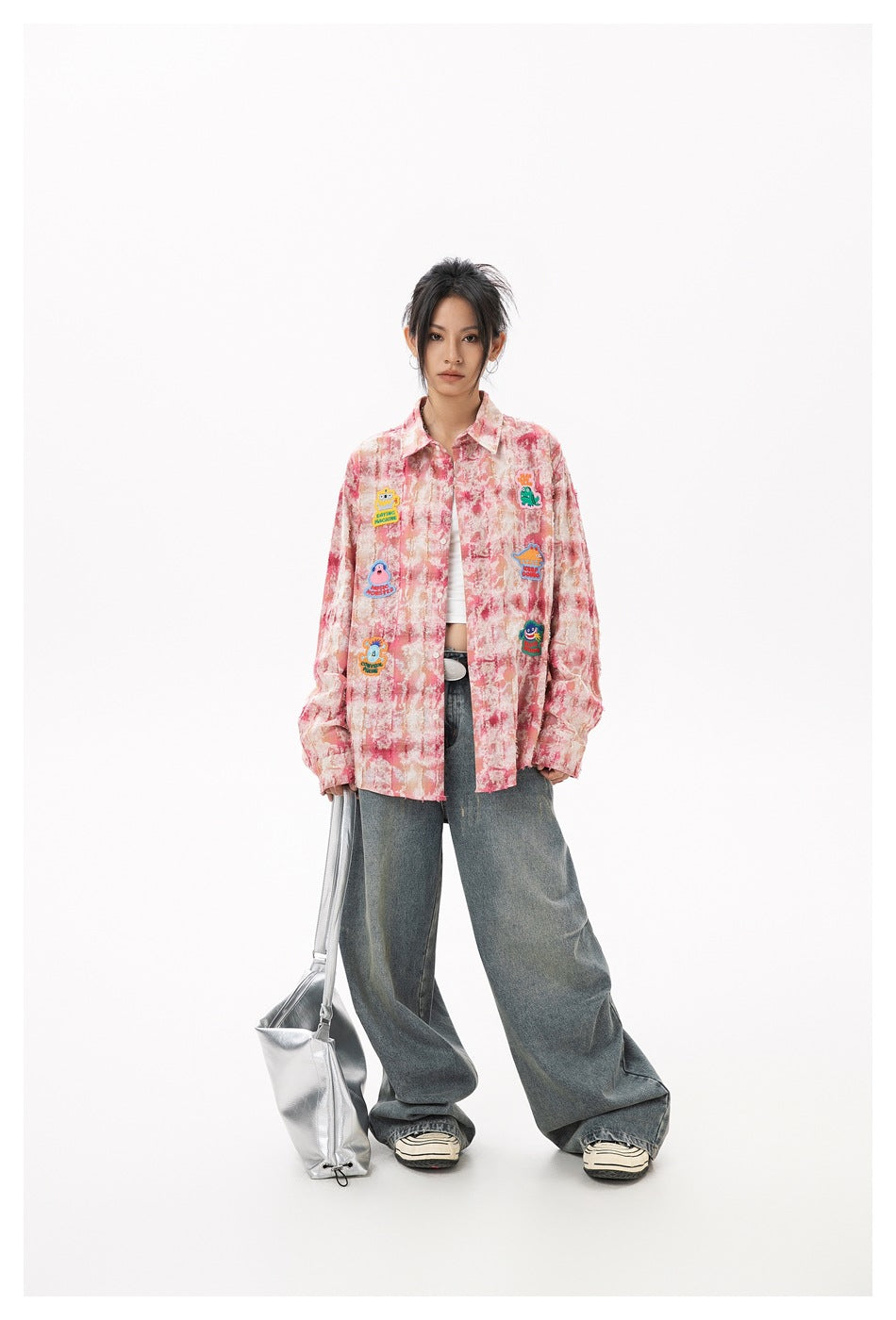 Full-length model wearing a pink marbled button-up shirt with colorful patches and wide-leg jeans, carrying a silver bag.