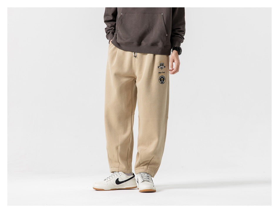 Front view of a model wearing beige joggers with a drawstring waist, ankle cuffs and small embroidered leg graphics.