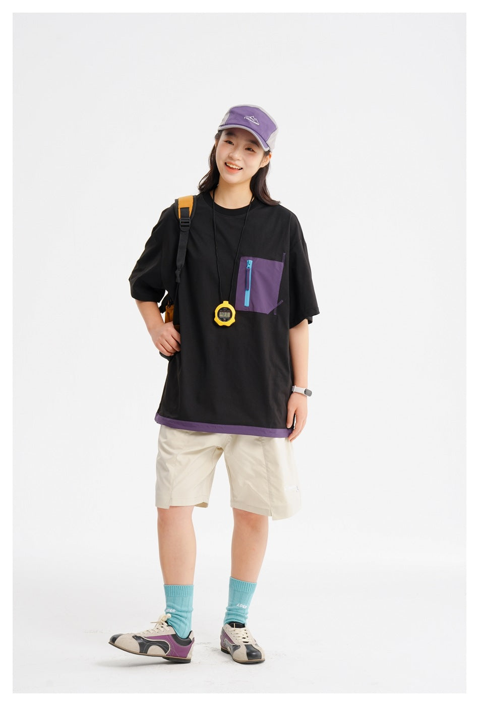 Model wearing a black oversized T-shirt with a purple zip chest pocket and hem trim, and beige shorts.