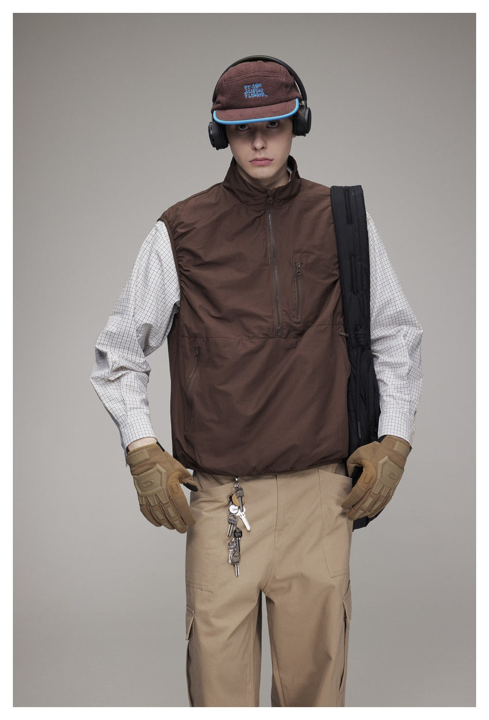 Model wearing a dark brown zip-front vest over a checked shirt, with beige cargo pants and a cap.