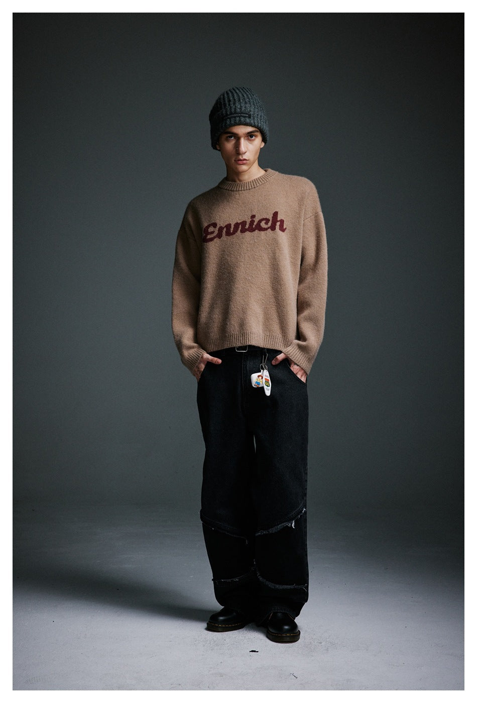 Model in a tan sweater with burgundy script lettering, loose black jeans, and a dark ribbed beanie, standing at a slight