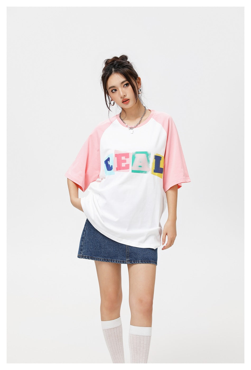 Model wearing a white T-shirt with pink raglan sleeves and colorful REAL lettering, shown with a denim skirt.