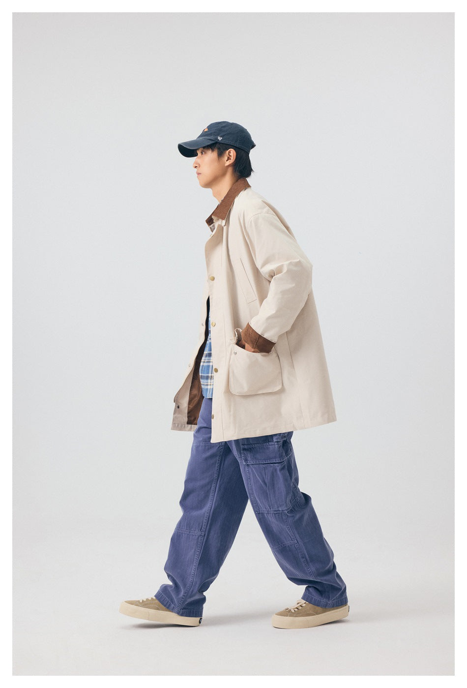 Side view of a model walking in a light beige coat with brown collar and cuffs, blue plaid shirt, and navy cargo pants.