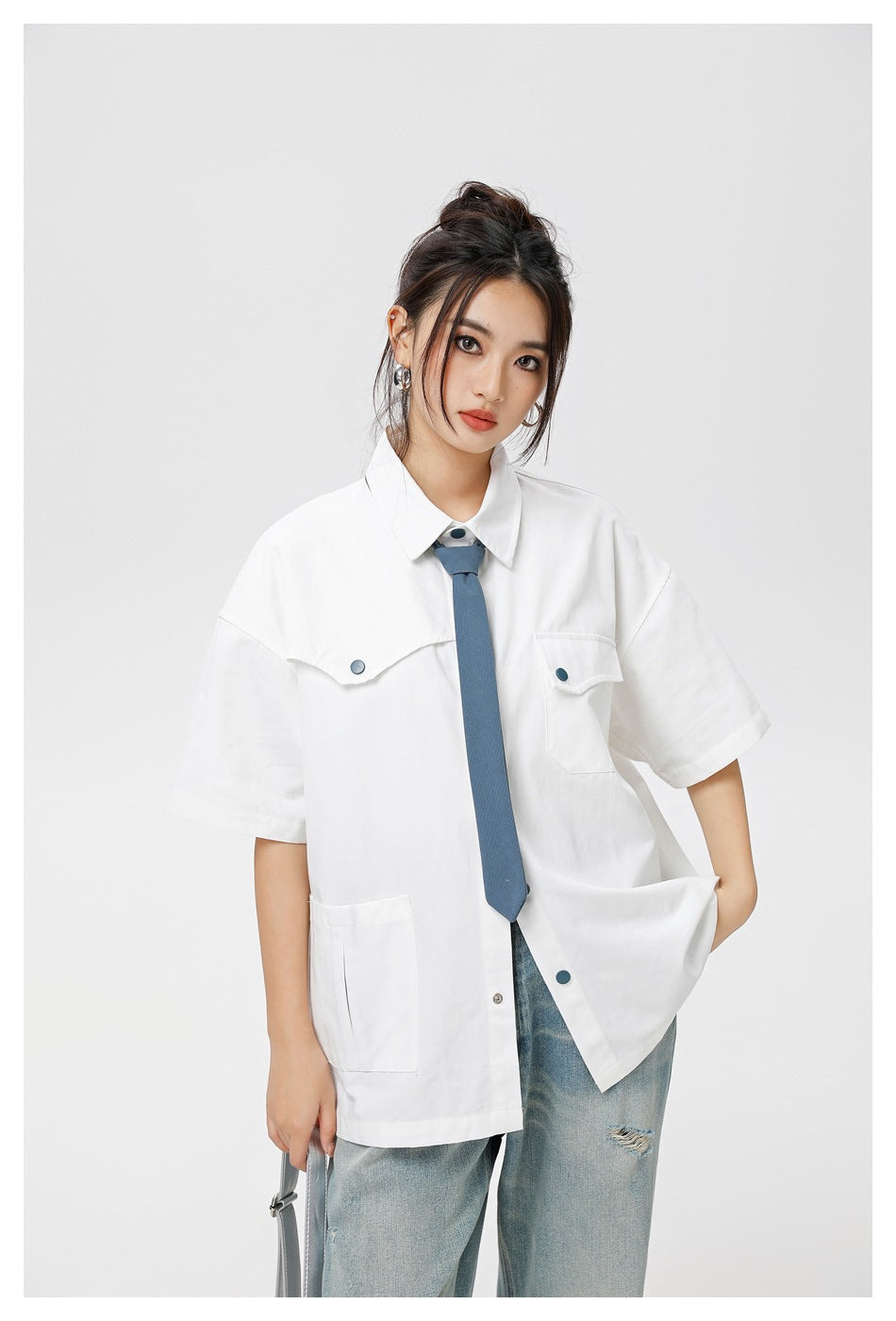 Model wearing an oversized white short-sleeve shirt with teal buttons and a blue tie, paired with light-wash jeans.