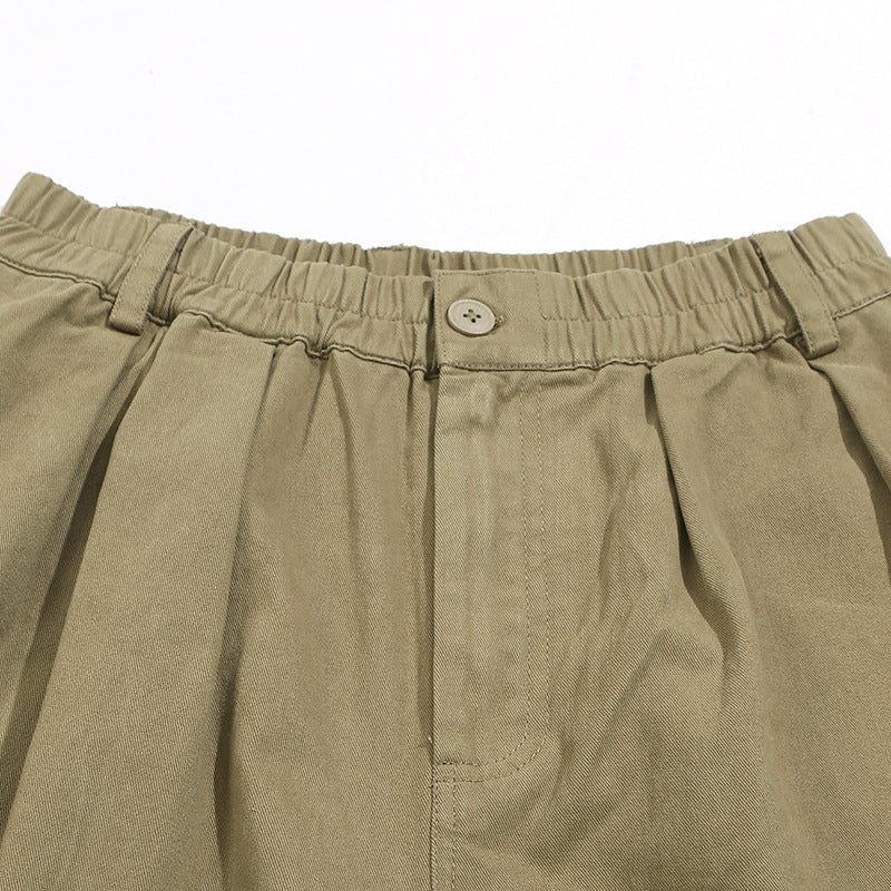 Close-up of khaki trousers showing the elastic waistband, button, belt loops, pleats and front fly.