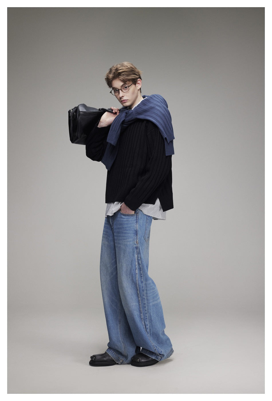 Model in a black ribbed sweater, checked shirt and wide-leg jeans, carrying a black bag with a blue sweater draped over his