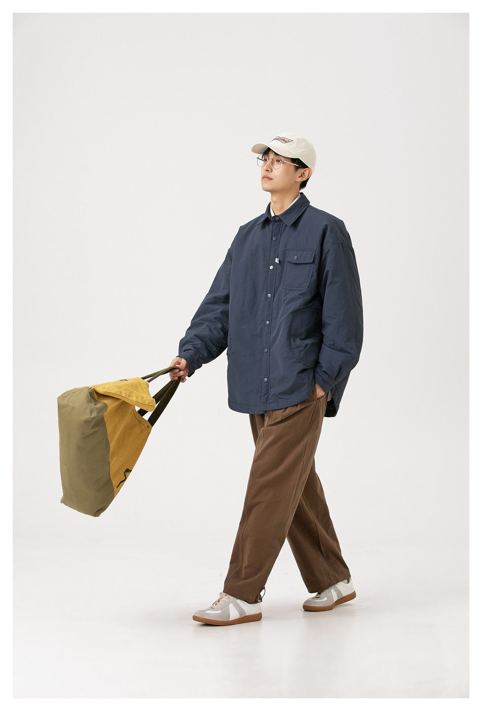 Full-length model walking in a navy snap-front jacket with a chest pocket, paired with brown trousers and a yellow tote.
