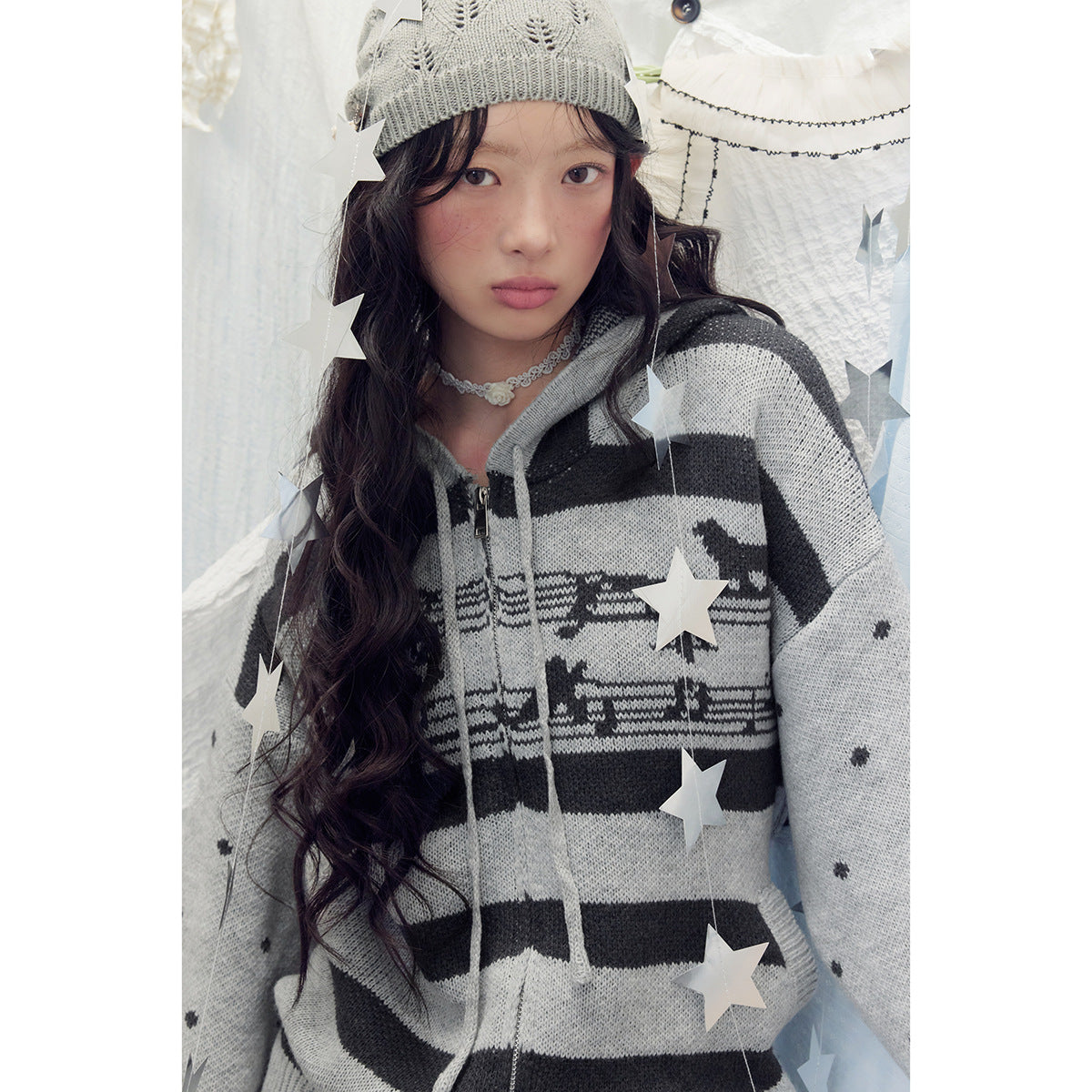 Close-up of a model wearing a gray zip-up hooded sweater with black stripes, a patterned chest band and dotted sleeves.